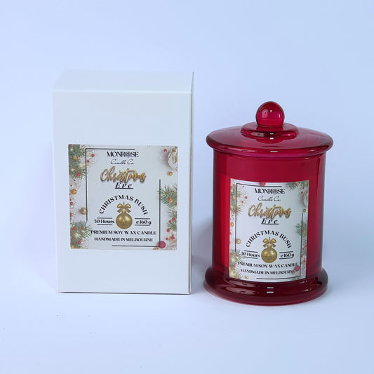 Front view of a red jar candle with 
"Christmas Bush" fragrance and a cotton wick, containing 160 grams of soy wax and offering 30 hours of burn time.